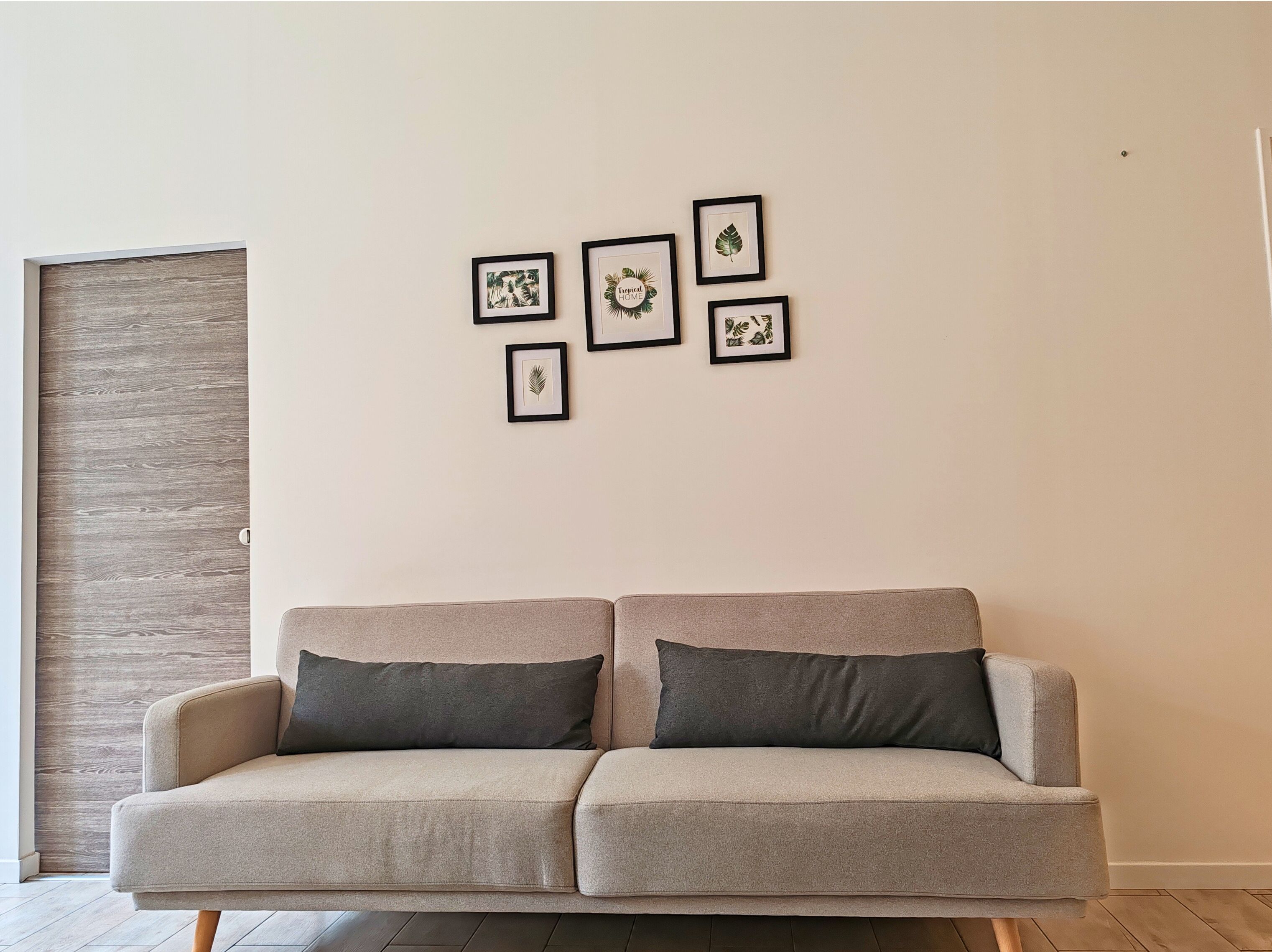 A neutral-colored sofa with dark cushions in a room with a plain wall and a wooden panel.