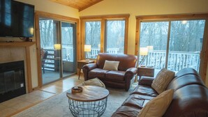A cozy living room with a fireplace, leather sofas, a coffee table, and large windows offering a view of a deck and trees.