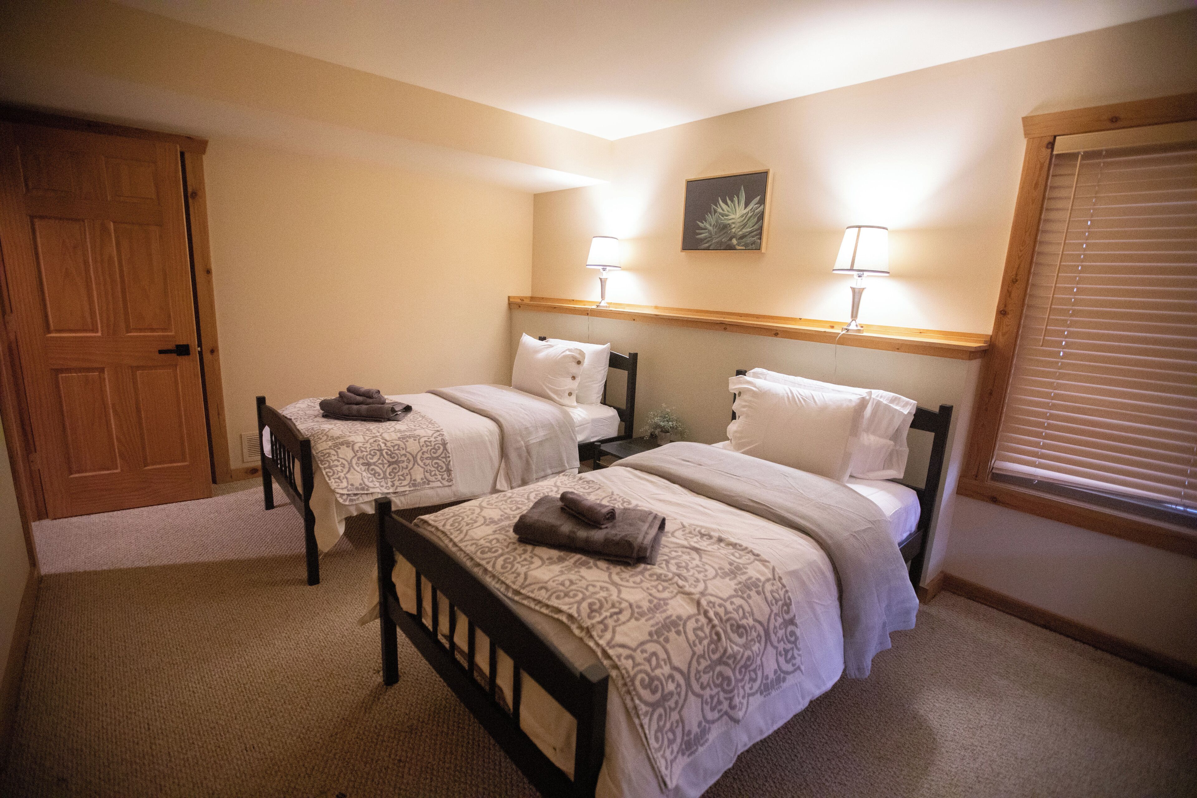 A hotel room with two beds, a wooden door, a small table, and a window with blinds.