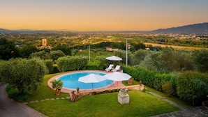 A swimming pool with lounge chairs and umbrellas, surrounded by lush greenery and a view of a distant town and mountains. - Villa Lontana in Lucca (Lucca)