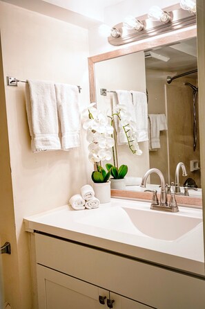 A bathroom with a white sink, a mirror, and a shower. There are white towels and a potted orchid on the counter.