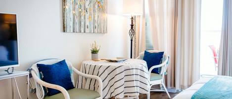 A hotel room with a bed, two white wicker chairs with blue cushions, a small table with a lamp, a television, and a painting on the wall.