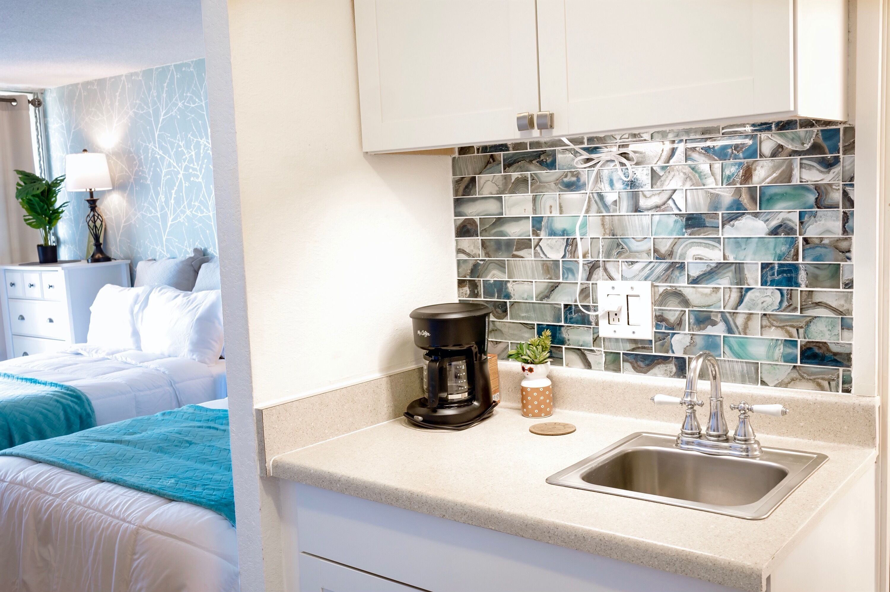 A hotel room with a bed, a coffee maker, a sink, and a decorative backsplash.