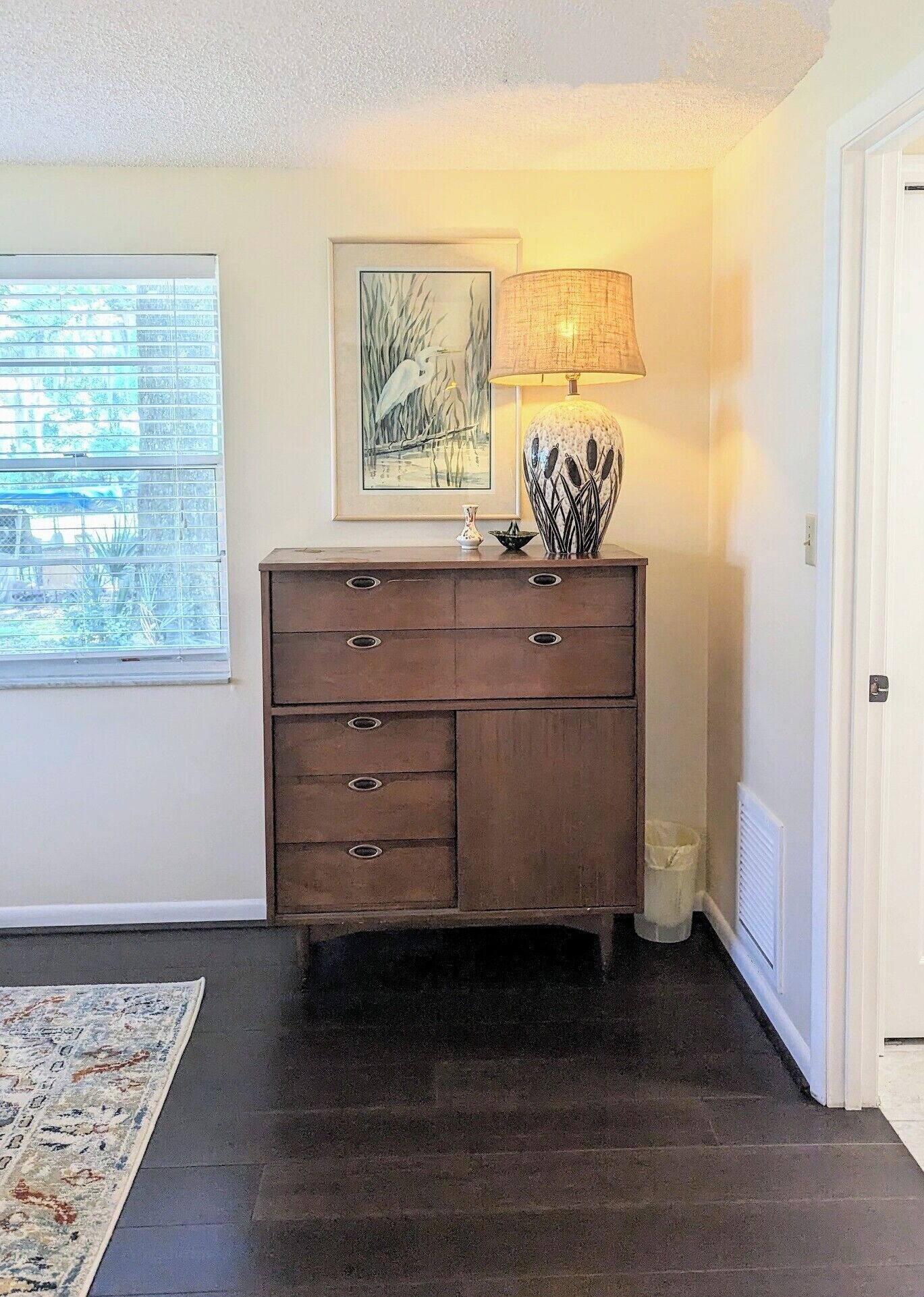 Une pièce avec une commode en bois, une lampe, un tableau encadré et une fenêtre avec des stores.