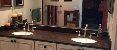 A bathroom with two sinks, a mirror, a clock, and a television mounted on the wall.