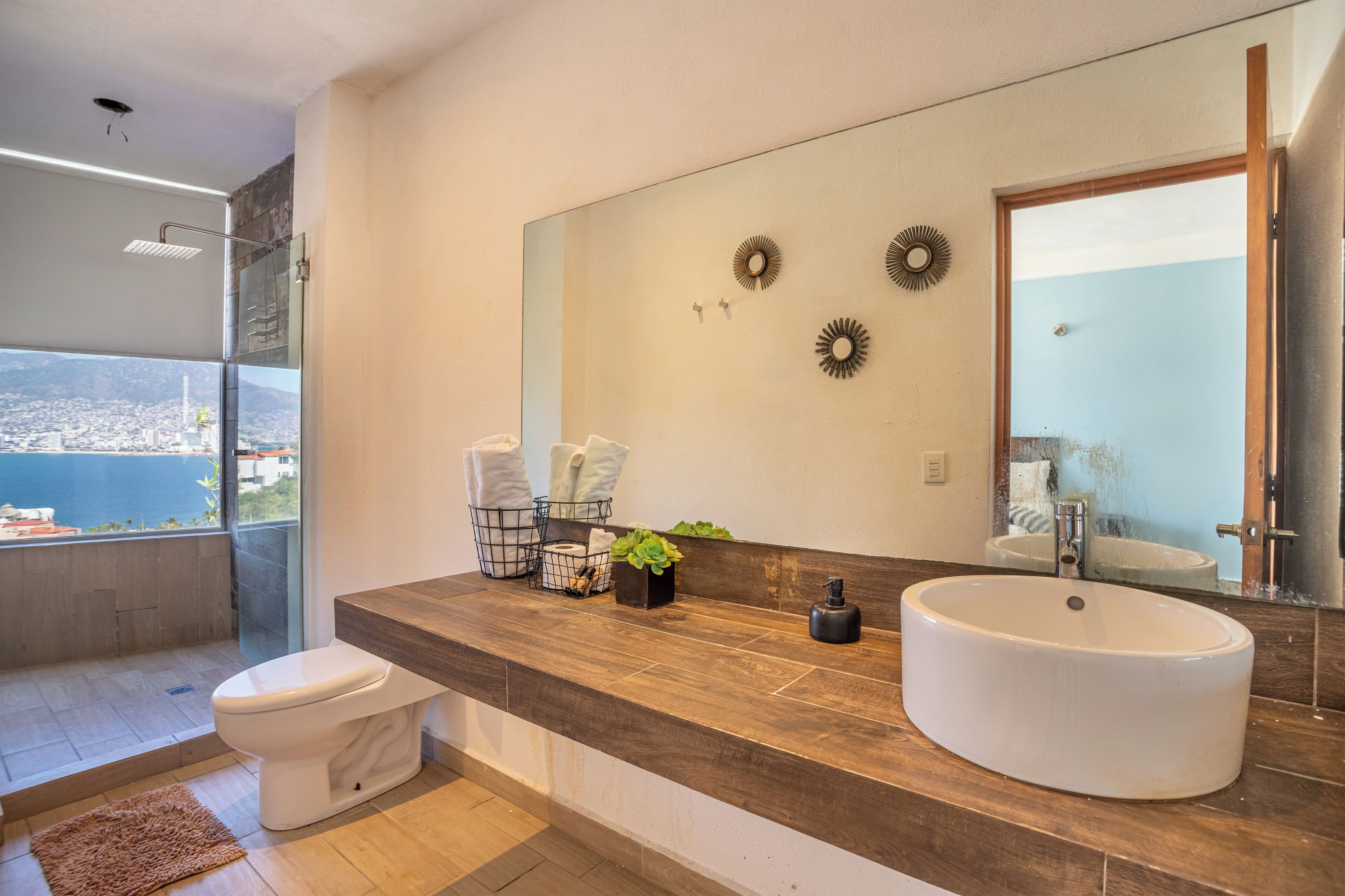 Ein modernes Badezimmer mit einem großen Spiegel, einem weißen Waschbecken und einer Toilette. Durch ein großes Fenster ist ein Blick auf die Stadt möglich.