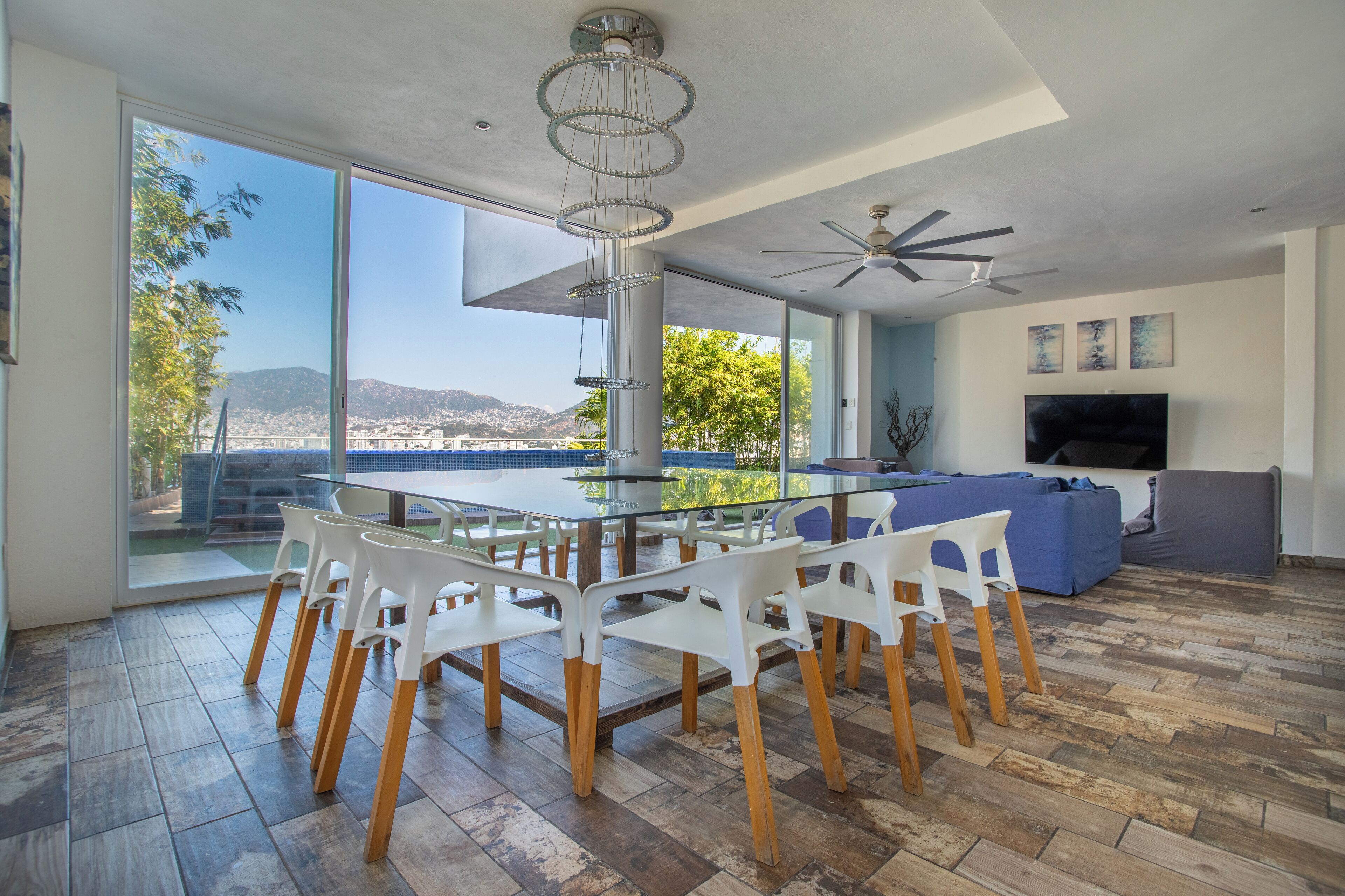 A modern living room with a glass dining table, white chairs, a blue sofa, and a TV.