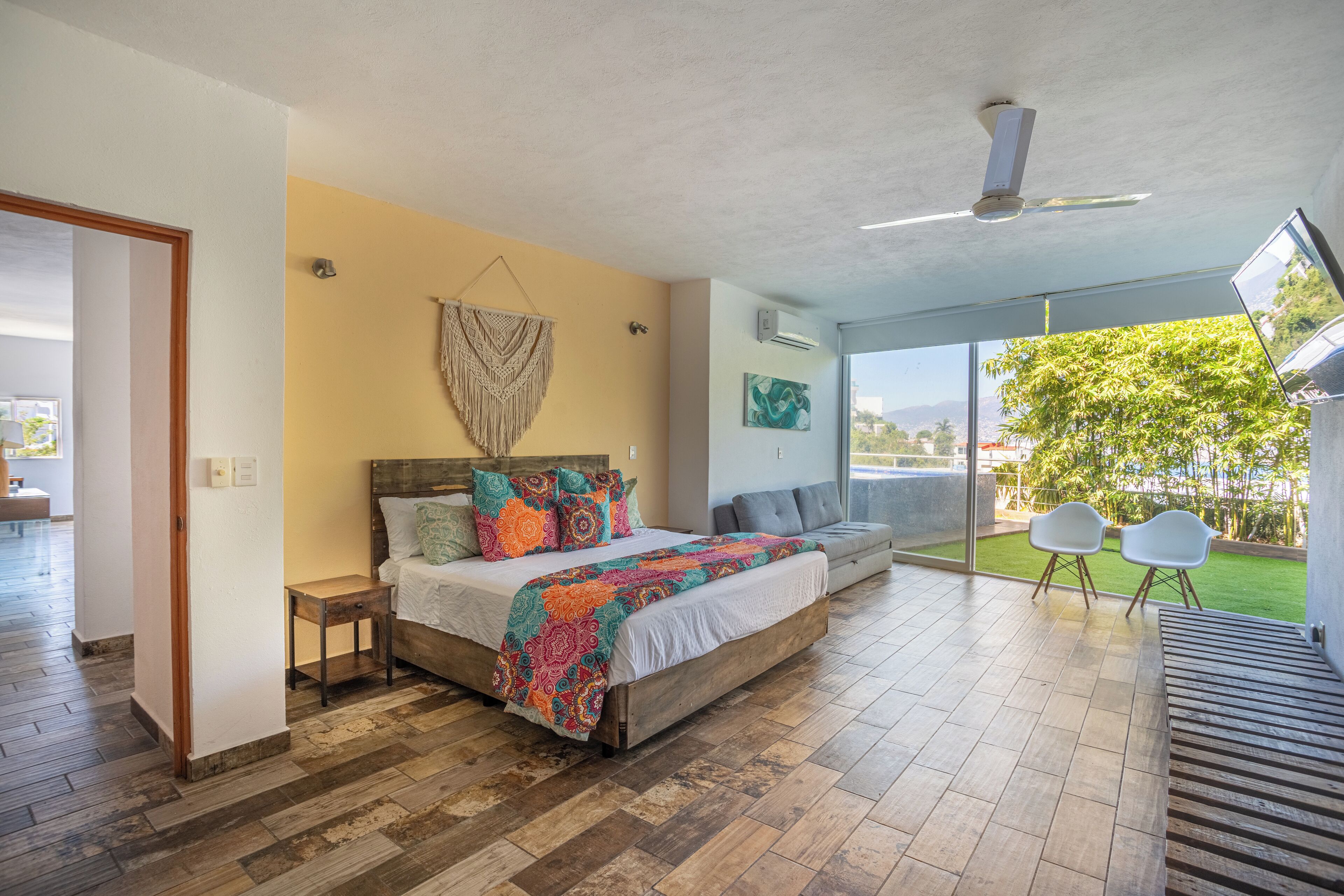 A bedroom with a large bed, a ceiling fan, a view of the outdoors, and a seating area with two chairs.