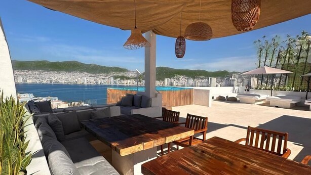 An outdoor seating area with a wooden table and cushioned seating, overlooking a cityscape and the sea. - LUXO Villa Acapulco Zona Brisas Guitarrón (Acapulco)