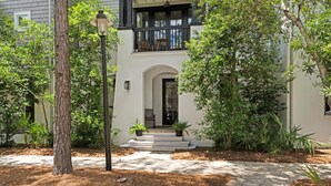 Une maison à deux étages, avec une façade blanche, un balcon noir et une entrée pavée avec des marches menant à une porte.