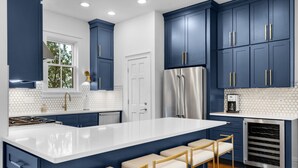 Une cuisine moderne avec des meubles de couleur bleu foncé, un plan de travail blanc et un réfrigérateur en acier inoxydable. On y trouve trois tabourets de bar avec des coussins beige et des pieds dorés.