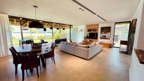 A modern living room with a sofa, dining table, and chairs, a TV, and a large window with a view. - Villa de Luxe La Paillotte (Black River)