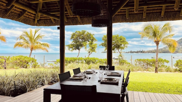 An outdoor dining area with a table set for two, overlooking a body of water with boats and palm trees. - Villa de Luxe La Paillotte (Black River)