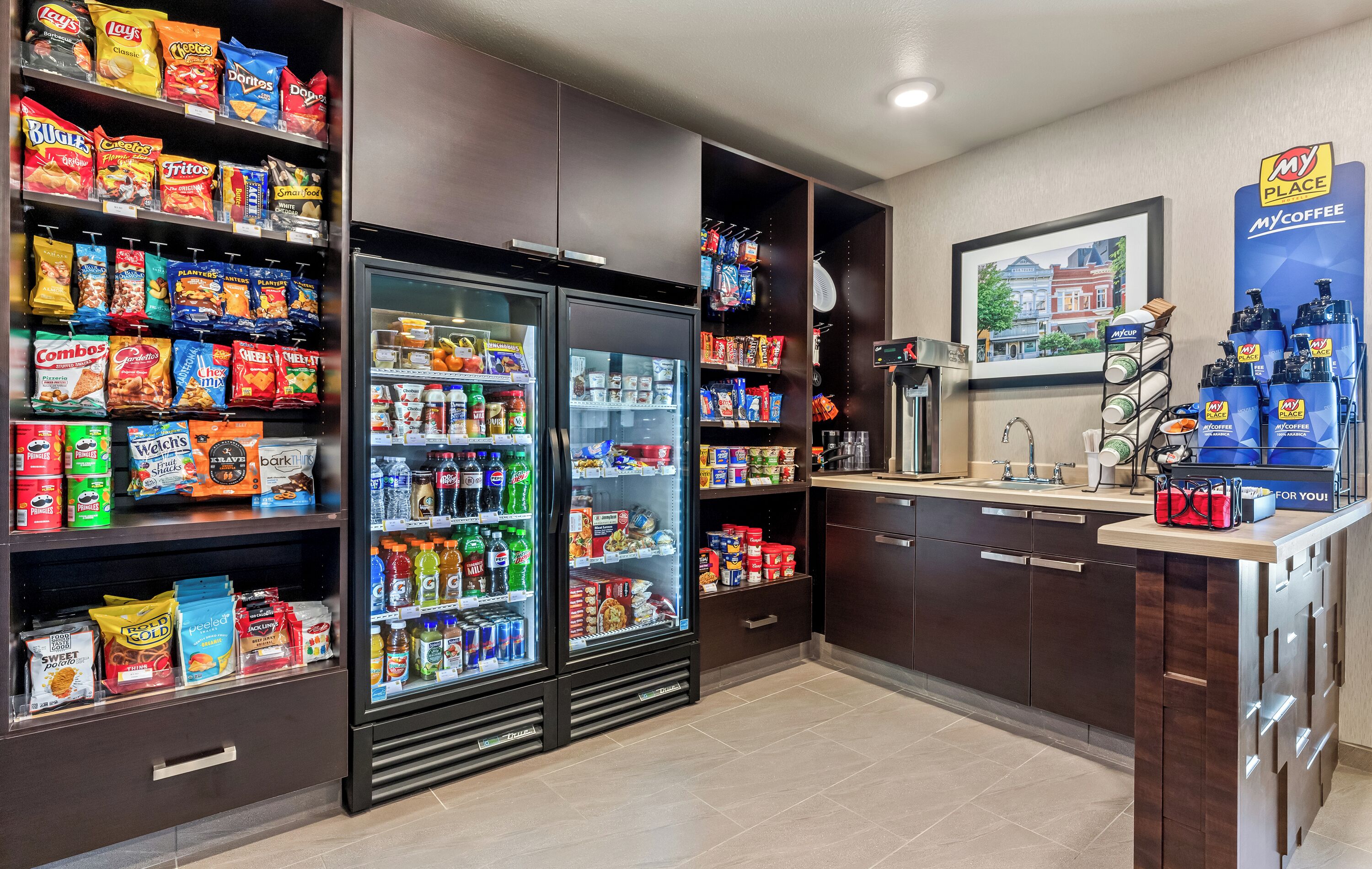A hotel kitchenette with shelves stocked with snacks, a refrigerator, and a coffee station.