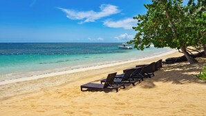 Een strand met helder turquoise water, een witte boot in de verte en ligbedden onder een palmboom.