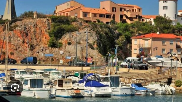 Un puerto deportivo con barcos amarrados, una ladera rocosa y edificios con tejados rojos al fondo.