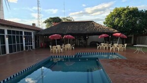 En swimmingpool med fliseterrasse, omgivet af udendørs siddepladser med parasoller.