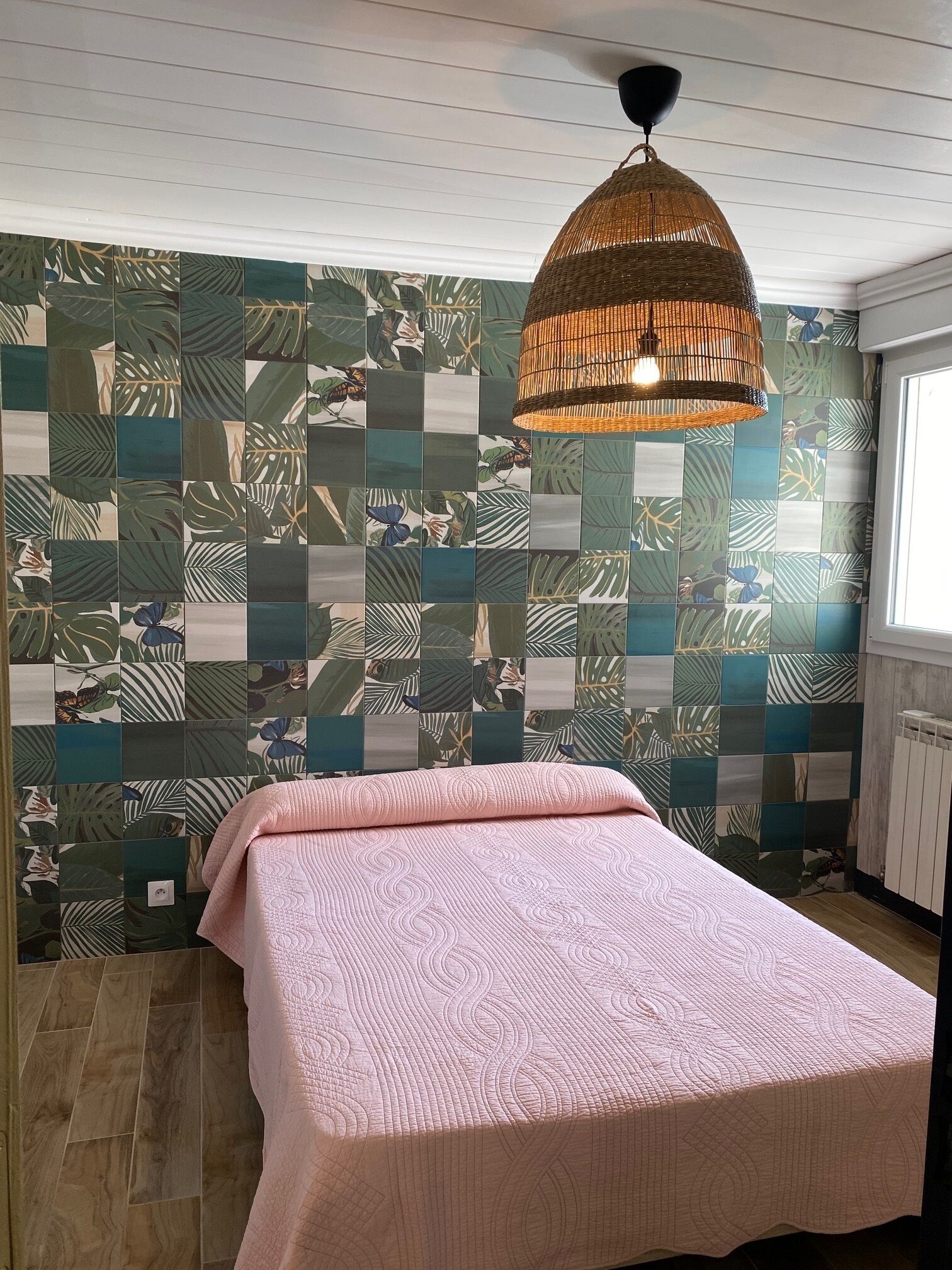 A bedroom with a bed, a hanging wicker lamp, and a wall with a tropical leaf pattern.