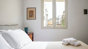 A neatly made bed with white linens, a small bedside table with a lamp, a framed picture on the wall, and a window with a view of buildings outside. - Casa Keller con Terrazza by Wonderful Italy (Noto)