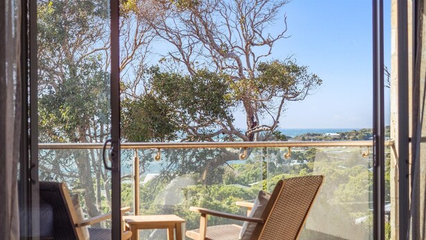 Un balcon avec deux fauteuils en osier et une petite table, offrant une vue panoramique sur un paysage verdoyant, avec des arbres et la mer à l’horizon.