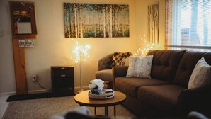 A cozy living room with a brown sofa, a small round coffee table with a decorative bowl, a wall-mounted shelf with a calendar, and a painting above the sofa. - Raven's Perch Cabin B Peaceful Getaway-Aurora-AlaskanCabin (Fairbanks)