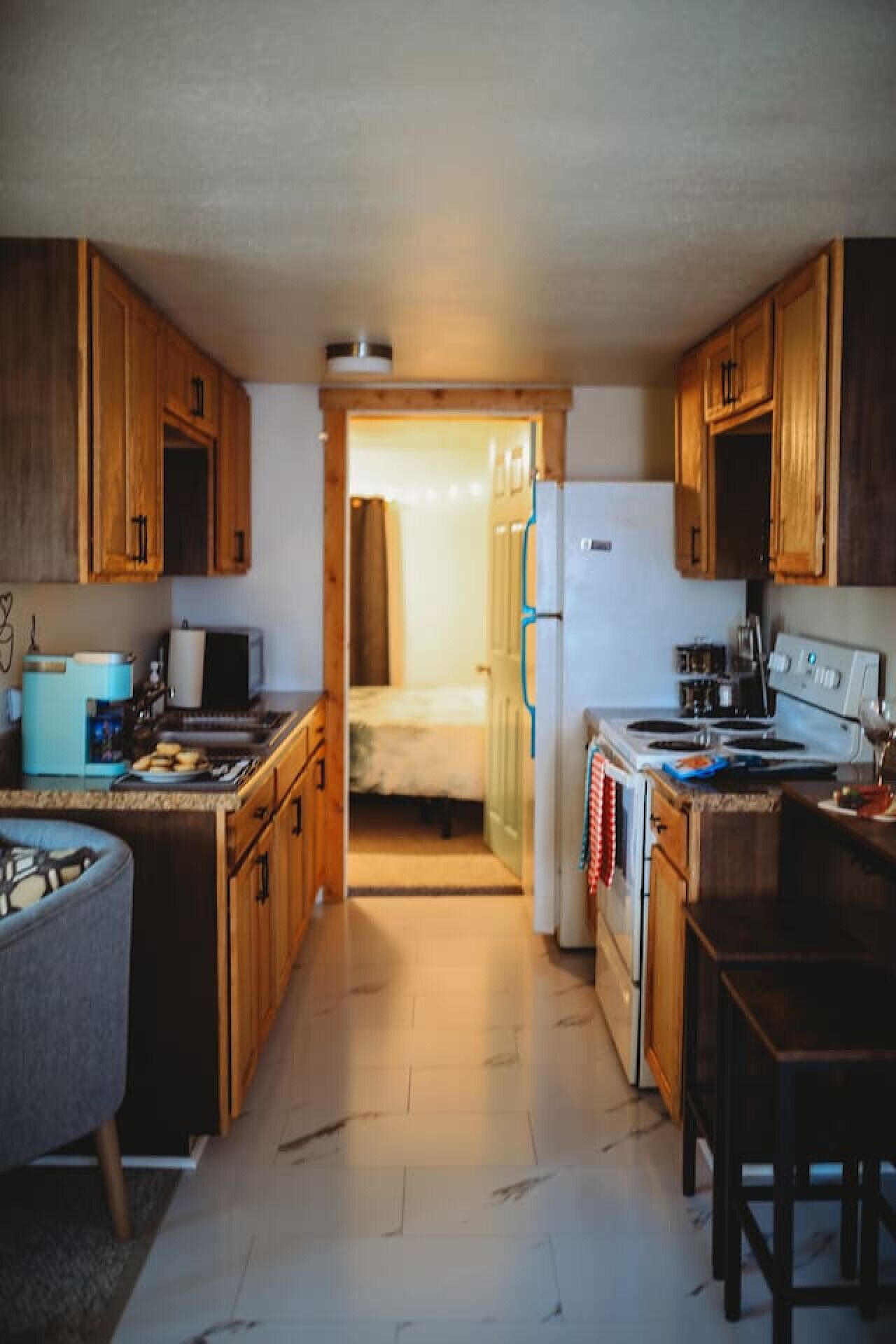 A compact kitchen with wooden cabinets, a refrigerator, a stove, and a sink. A bed is visible through an open doorway in the background.