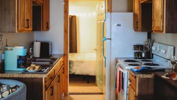 A compact kitchen with wooden cabinets, a refrigerator, a stove, and a sink. A bed is visible through an open doorway in the background.