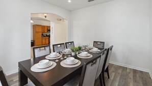 A dining area with a long table set for six, surrounded by chairs. A kitchen is visible in the background. - 5/3 Pool home located in Sunset Ridge Community! (Davenport)
