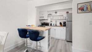 Een compacte keuken met witte kastjes, een magnetron, een fornuis en een koelkast. Er zijn twee blauwe barkrukken en een klein witte tafel.