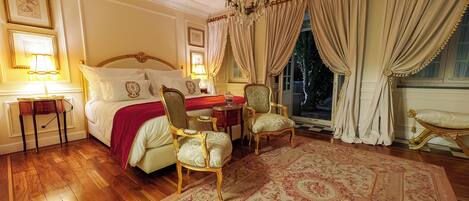 A bedroom with a large bed, two chairs, a chandelier, and a view of the outdoors.