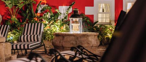 A cozy outdoor seating area with striped chairs, a stone planter with a fountain, and lush greenery.