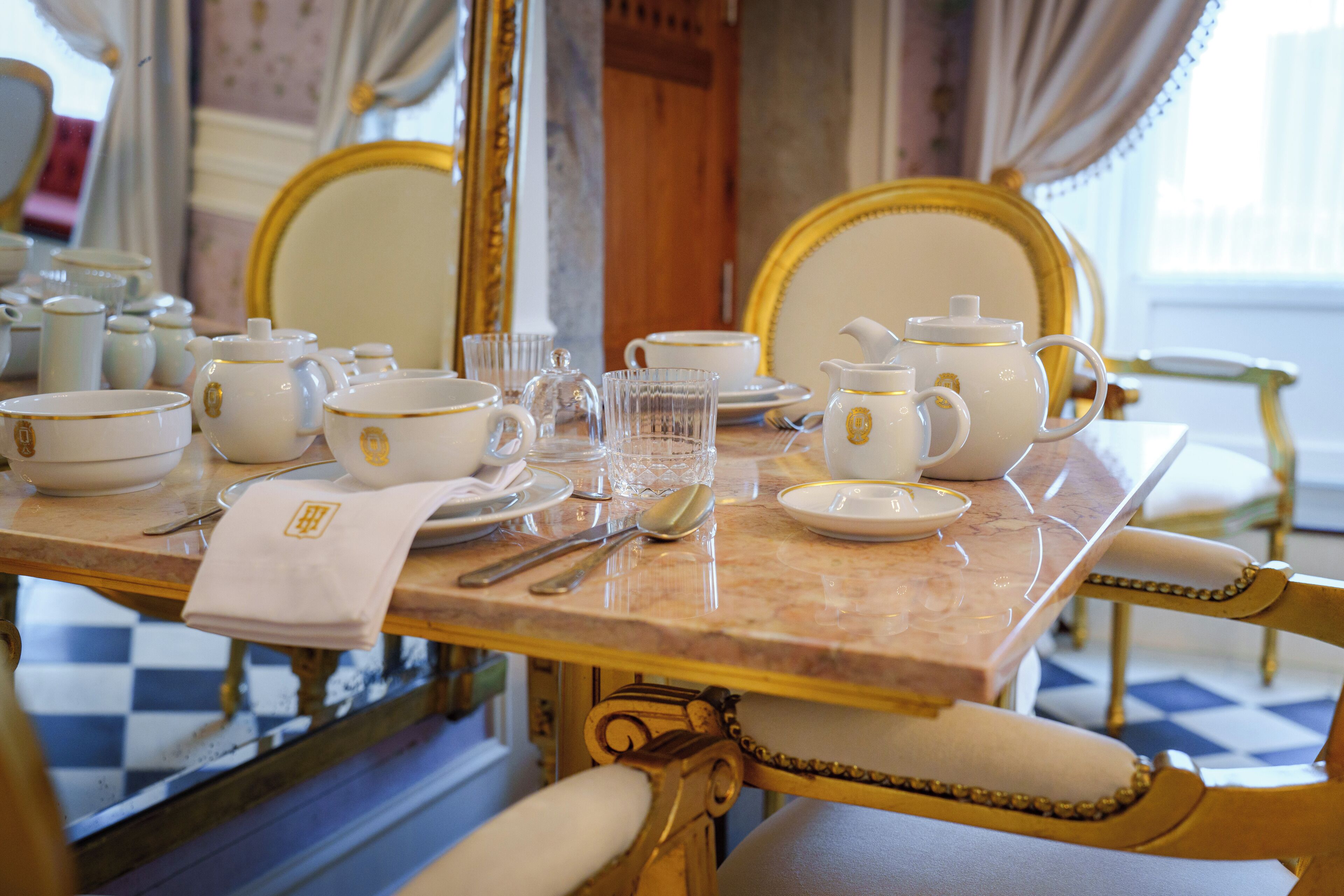 A dining table set with white and gold china, glassware, and silverware, surrounded by ornate chairs.