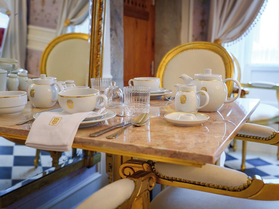A dining table set with white and gold china, glassware, and silverware, surrounded by ornate chairs.