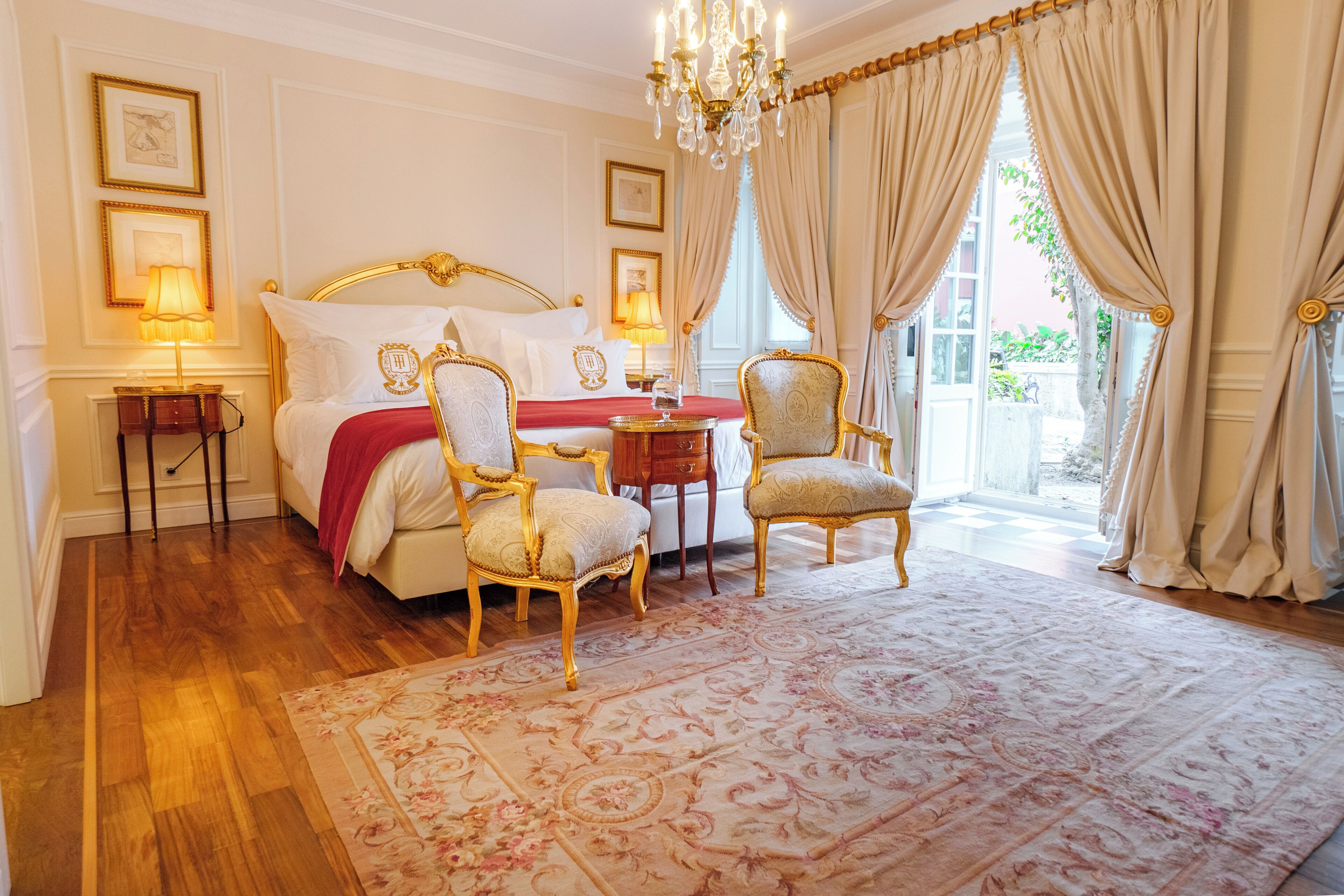 A luxurious bedroom with a large bed, ornate chairs, a chandelier, and a view of the outdoors.