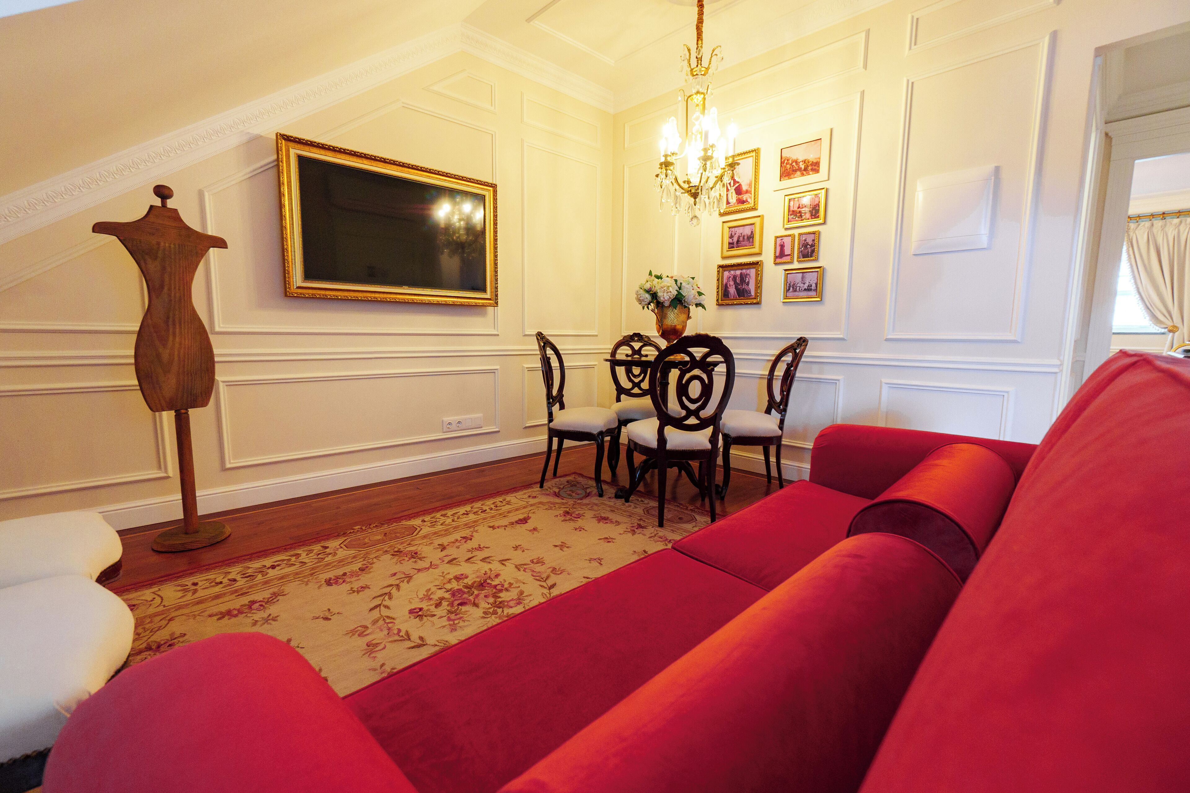 A room with a red sofa, a dining table with chairs, a chandelier, and a large framed picture on the wall.
