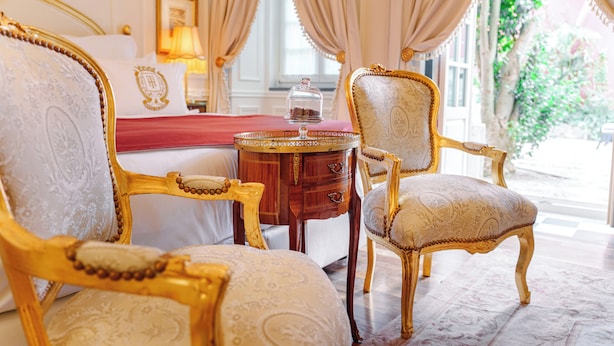 Un dormitorio lujoso con muebles ornamentados, colcha roja y vistas a la vegetación.