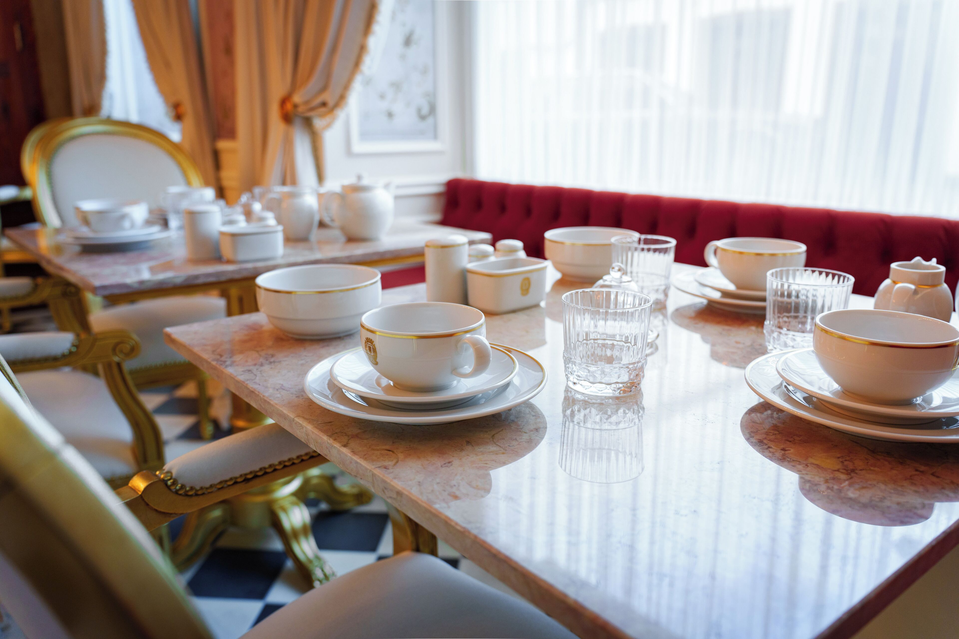 Una mesa de comedor con vajilla blanca y dorada, cristalería transparente y un banco tapizado en rojo.