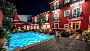 Una piscina con diseño de cuadros, rodeada por una terraza de madera y edificios rojos con balcones.