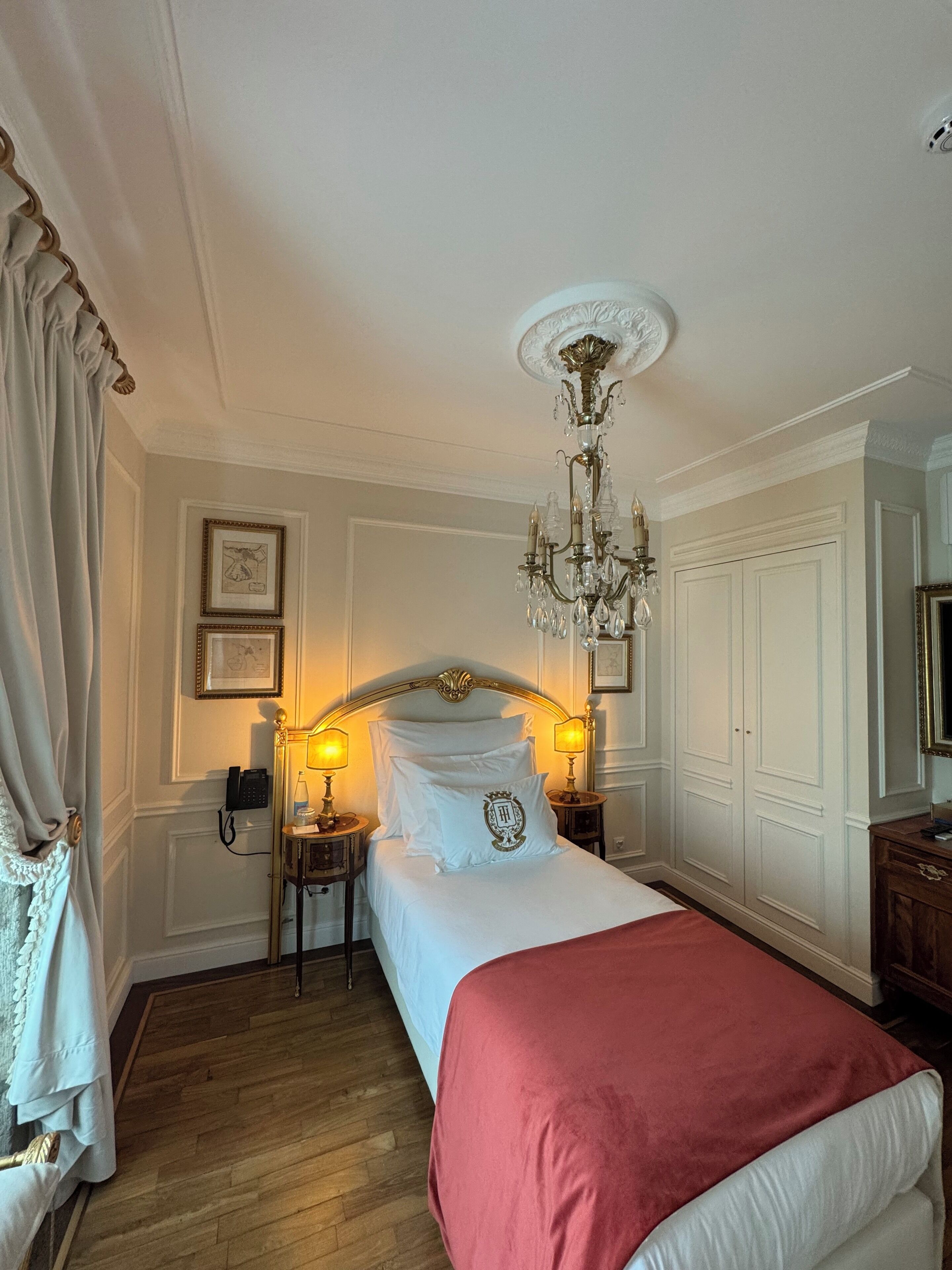A hotel room with a bed, a chandelier, a telephone, and framed pictures on the wall.