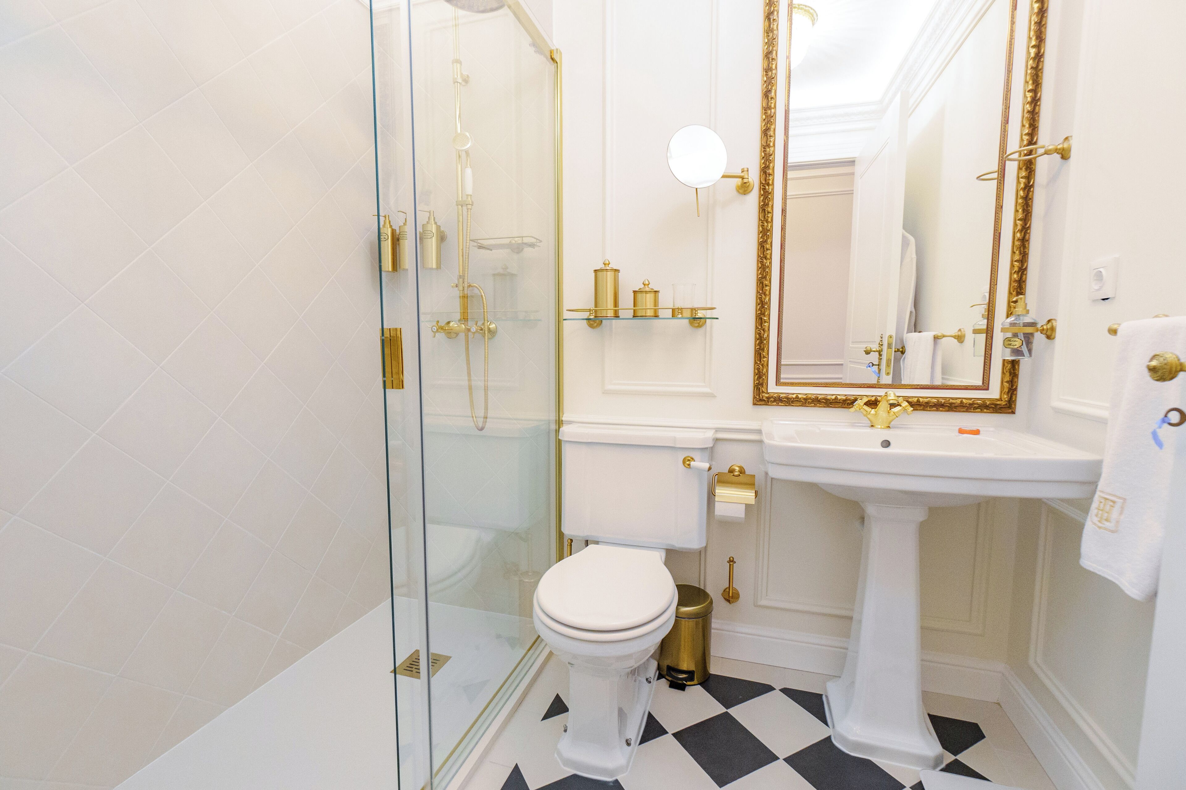 Une salle de bain avec une douche à parois de verre, des toilettes blanches, un lavabo sur colonne et un miroir avec un cadre doré.
