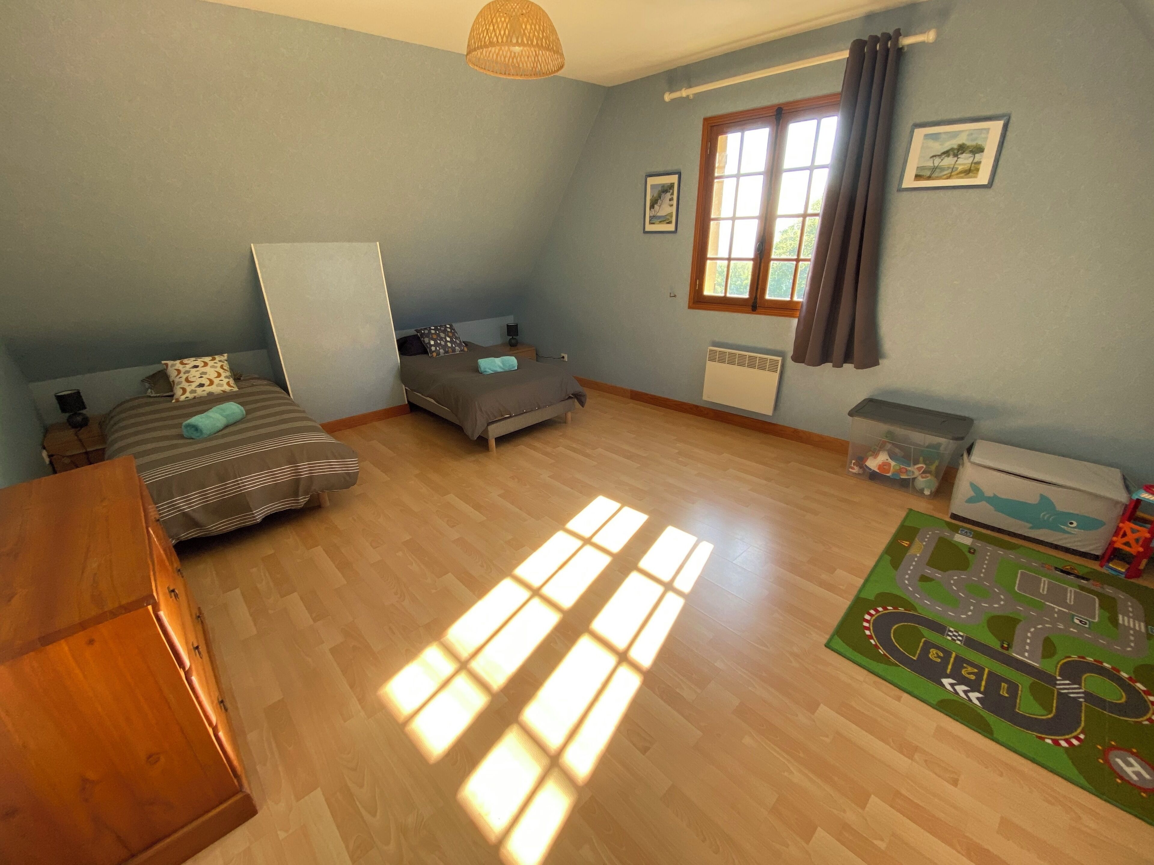 A room with two beds, a wooden wardrobe, a radiator, a window with curtains, and a green mat with a road design.