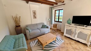 A living room with a sofa, a wooden coffee table, a wicker chair, a TV on a cabinet, and a window with curtains. - "Sur la colline": country house with heated pool, near Sarlat. (Domme)