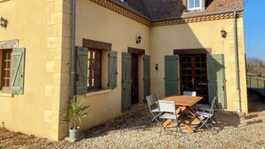 A yellow house with green shutters, a wooden table with chairs, and a gravel driveway. - "Sur la colline": country house with heated pool, near Sarlat. (Domme)