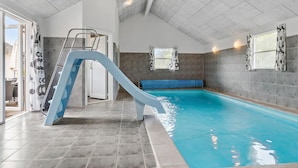 En inomhuspool med en blå vattenrutschkana, kaklade väggar och en bänk.