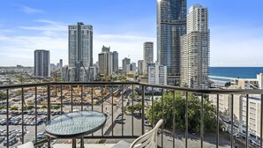 A balcony with a glass table and chairs overlooking a cityscape with tall buildings and a beach. - Twin Unit with Ocean View in Surfers Paradise (Surfers Paradise)