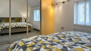A modern bedroom with a geometric-patterned bedspread, a wooden wardrobe, a bedside table with a lamp, and a window with blinds.