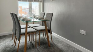 A dining area with a glass table and grey upholstered chairs, a grey wall with three framed paintings, and a window with blinds.