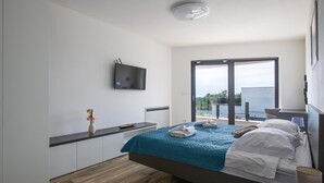 Une chambre moderne avec un lit, un téléviseur fixé au mur, un bureau et une grande fenêtre donnant sur l’extérieur.