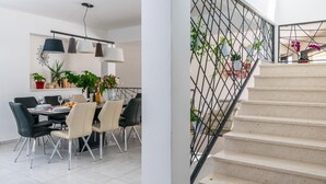 Un espace de salle à manger moderne, avec une table dressée pour quatre personnes, entourée de chaises. On aperçoit un escalier avec une rampe en métal et des plantes en pot.