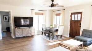 Una sala de estar con televisión de pantalla plana, piso de madera, una mesa de comedor con sillas, un sofá y una puerta con ventanas.